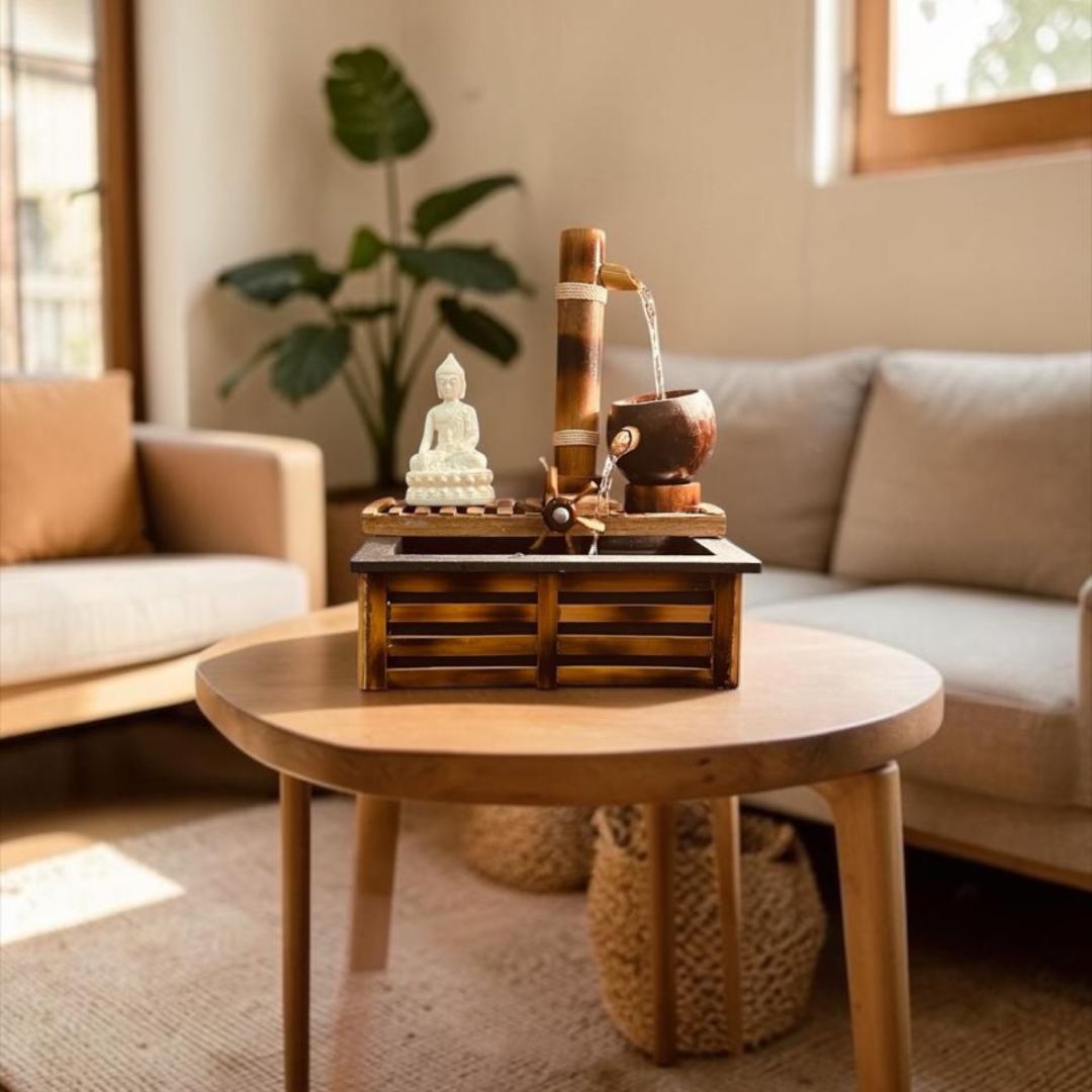 A round wooden coffee table in a cozy living room setting, styled with a bamboo and coconut water feature, a small white Buddha statue, and a decorative tray.